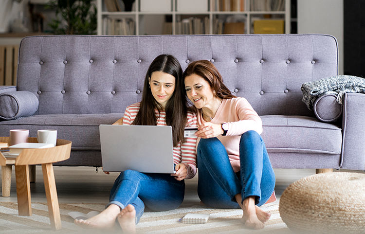 Parent and child sitting together on floor and looking at a laptop. Parent holds a credit card.